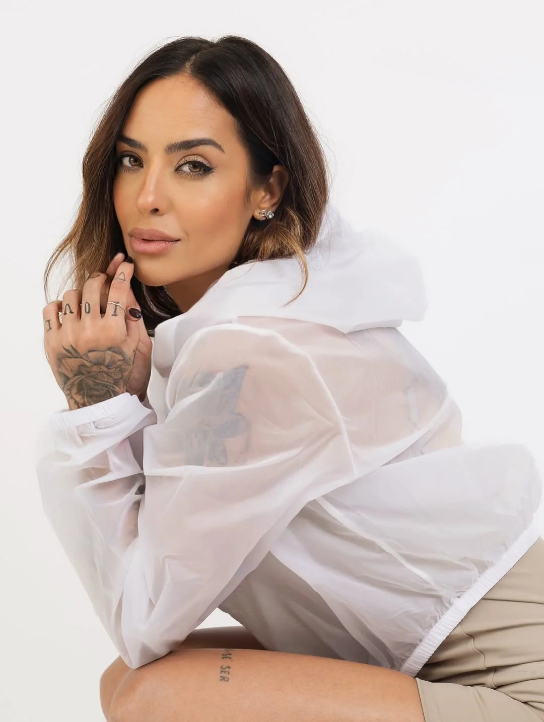 Julia Melo com jaqueta transparente branca, apoiada de lado e olhando para a câmera.
