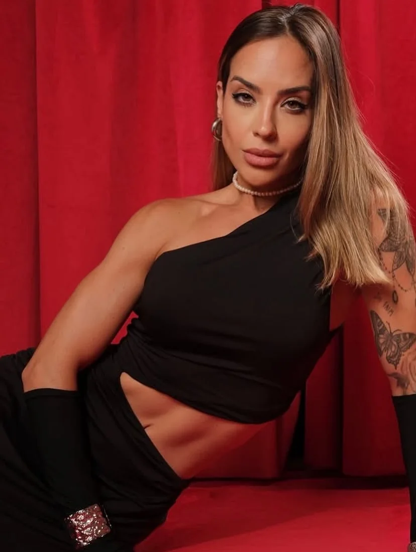 Julia Melo de conjunto preto de um ombro só, sentada diante de uma cortina vermelha.